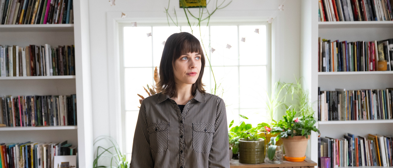 Paige Lewis looks off to the side as she stands in front of a window and two large bookshelves.
